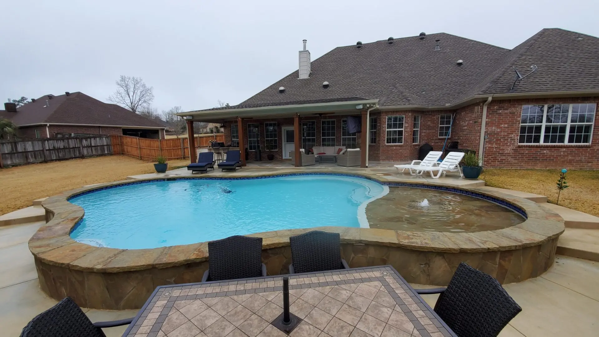 Freeform custom pool with a flagstone tanning ledge in front of a brick home with mature trees